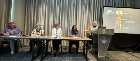 table with 4 white women sitting and one woman is standing behind a podium